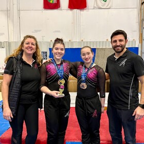 Deux athlètes en justaucorps noir et rose posent avec des médailles et un trophée aux côtés de deux entraîneurs souriants, devant trois drapeaux canadiens suspendus.